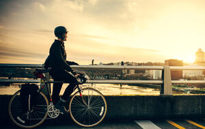 Radfahrerin mit Helm steht auf Weg mit Fahrrad und blickt auf einen Fluss bei Sonnenuntergang.