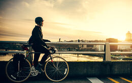 Radfahrerin mit Helm steht auf Weg mit Fahrrad und blickt auf einen Fluss bei Sonnenuntergang.
