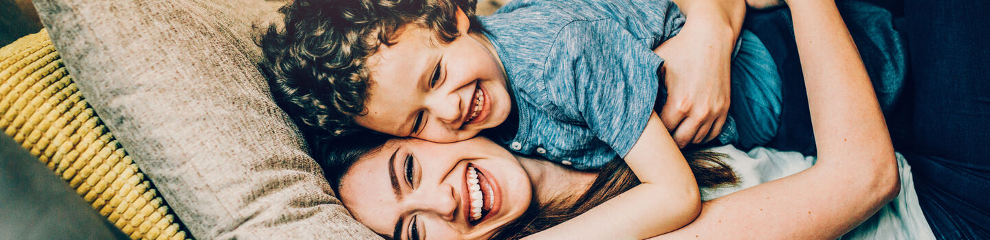 Eine junge Frau kuschelt mir ihrem kleinen Kind am Sofa, beide lachen. Das Kind liegt auf der Mutter und wird von ihr liebevoll festgehalten. 