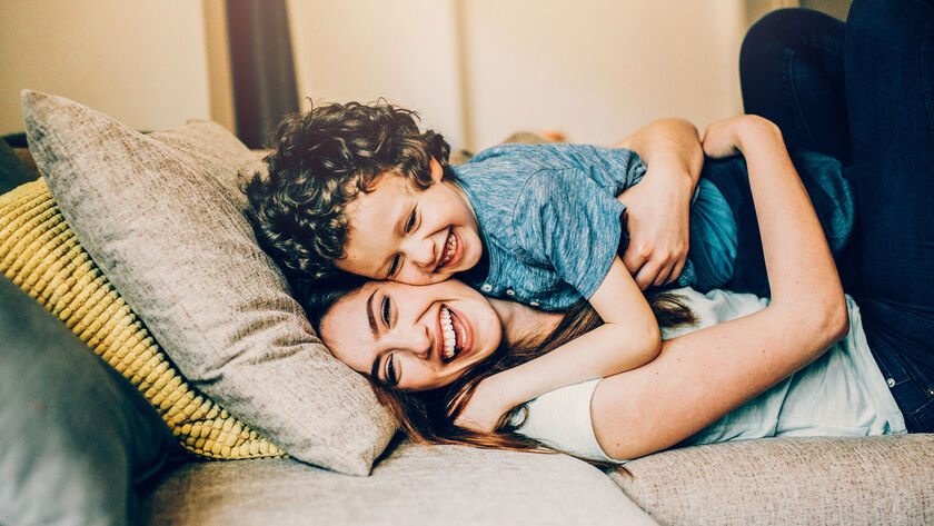 Eine junge Frau kuschelt mir ihrem kleinen Kind am Sofa, beide lachen. Das Kind liegt auf der Mutter und wird von ihr liebevoll festgehalten.