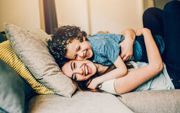 Eine junge Frau kuschelt mir ihrem kleinen Kind am Sofa, beide lachen. Das Kind liegt auf der Mutter und wird von ihr liebevoll festgehalten.