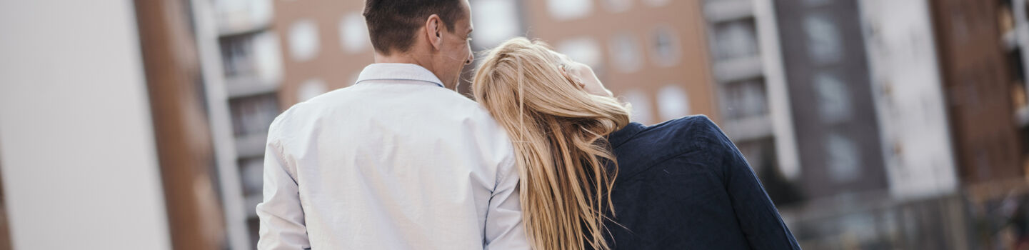 Eine Frau und ein Mann gehen zwischen einem Häuserblock und halten Hände. Das Pärchen ist von hinten zu sehen und die Frau lehnt ihren Kopf an die Schulter des Mannes.