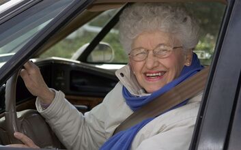Ältere Frau mit weißem Haar und Brille lachend am Steuer eines Autos.