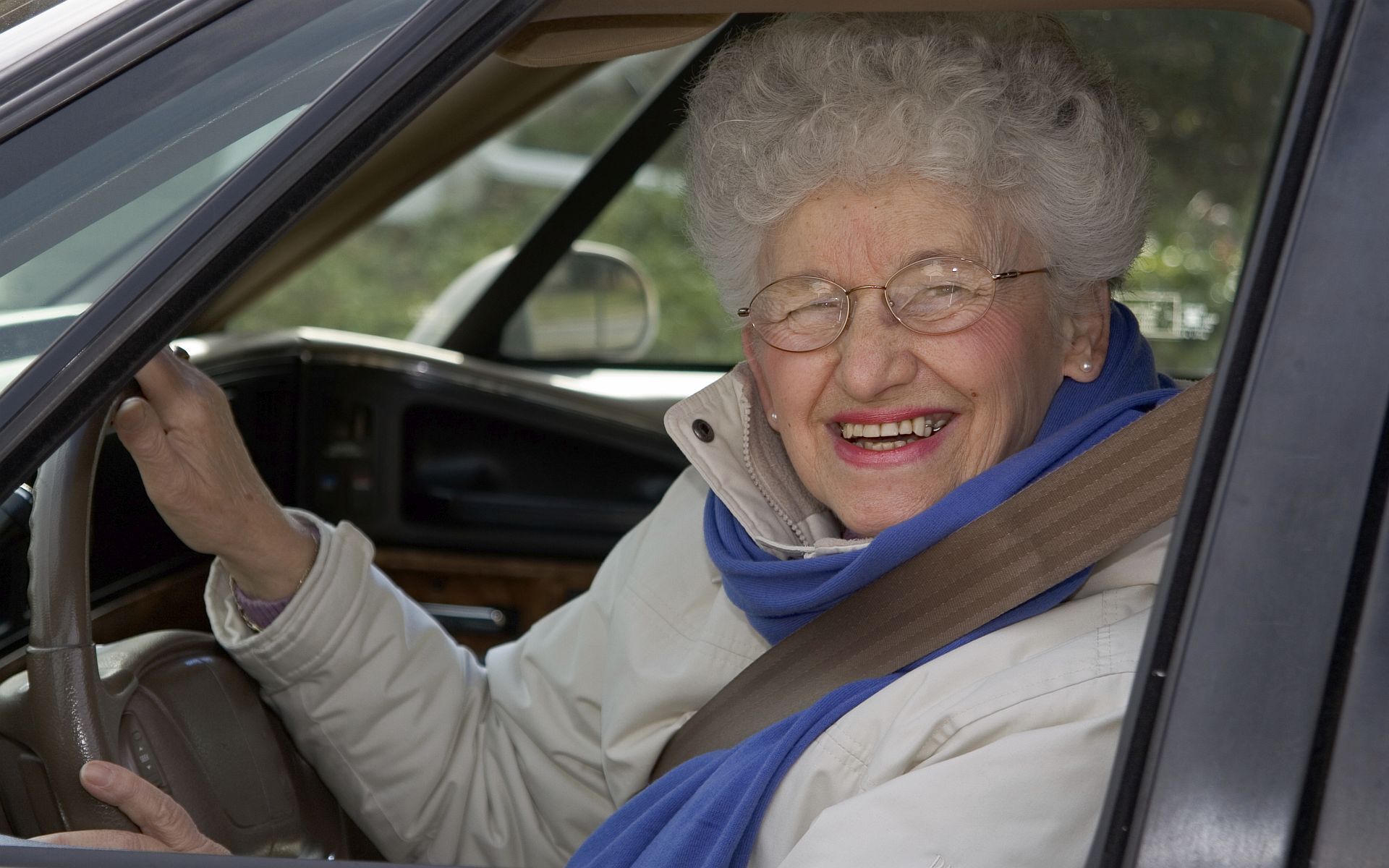 Ältere Frau mit weißem Haar und Brille lachend am Steuer eines Autos.