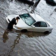 Überschwemmte Straße mit einem Auto. Das Auto ist bis zu den Reifen im Wasser. Ein Mann steht beim Auto und telefoniert.