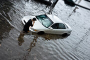 Überschwemmte Straße mit einem Auto. Das Auto ist bis zu den Reifen im Wasser. Ein Mann steht beim Auto und telefoniert.