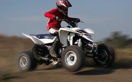 Eine Person fährt auf einem weißen Quad, auch Trike genannt, im Gelände auf einem Hügel. Sie trägt einen Motorradhelm, Stiefel, einen roten Sweater und eine weiße Hose.