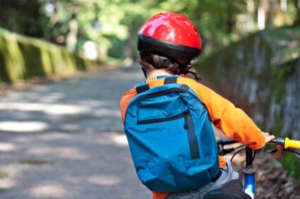 Ein Kind fährt auf einem Fahrrad eine Straße entlang. Man sieht es von hinten. Es trägt einen roten Fahrradhelm und eine blaue Schultasche am Rücken.