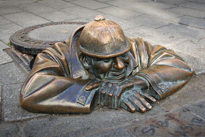 eine Bronzefigur, die aus einem Kanaldeckel schaut