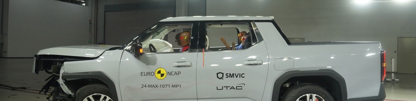 Crashtest von einem weißen großen Pickup. Die Motorhaube ist stark eingedrückt und kaputt. Das Fahrzeug steht in einer Testhalle.