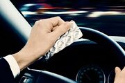 Das Bild zeigt die Hände von einem Autofahrer im Fahrzeug. Er sitzt am Steuer und hält mit einer Hand das Lenkrad, während er in der linken Hand ein Tablettenblister hält. 