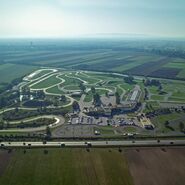Inmitten einer ländlichen Landschaft erstreckt sich das Fahrtechnik Zentrum Teesdorf. Es gibt viele asphaltierte Trainingsstrecken mit Kreuzungselementen und Kurvenkombinationen, einen Offroad Hügel mit einem Teich, einen Handlingparcours im Motorrad Zentrum, ein Gebäude und einen großen Parkplatz.  Im Hintergrund ist ein Dorf inmitten grüner Felder.  