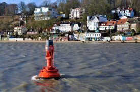 Eine rote Boje im Wasser in der Nähe eines Ufers mit Häusern und einem Strand