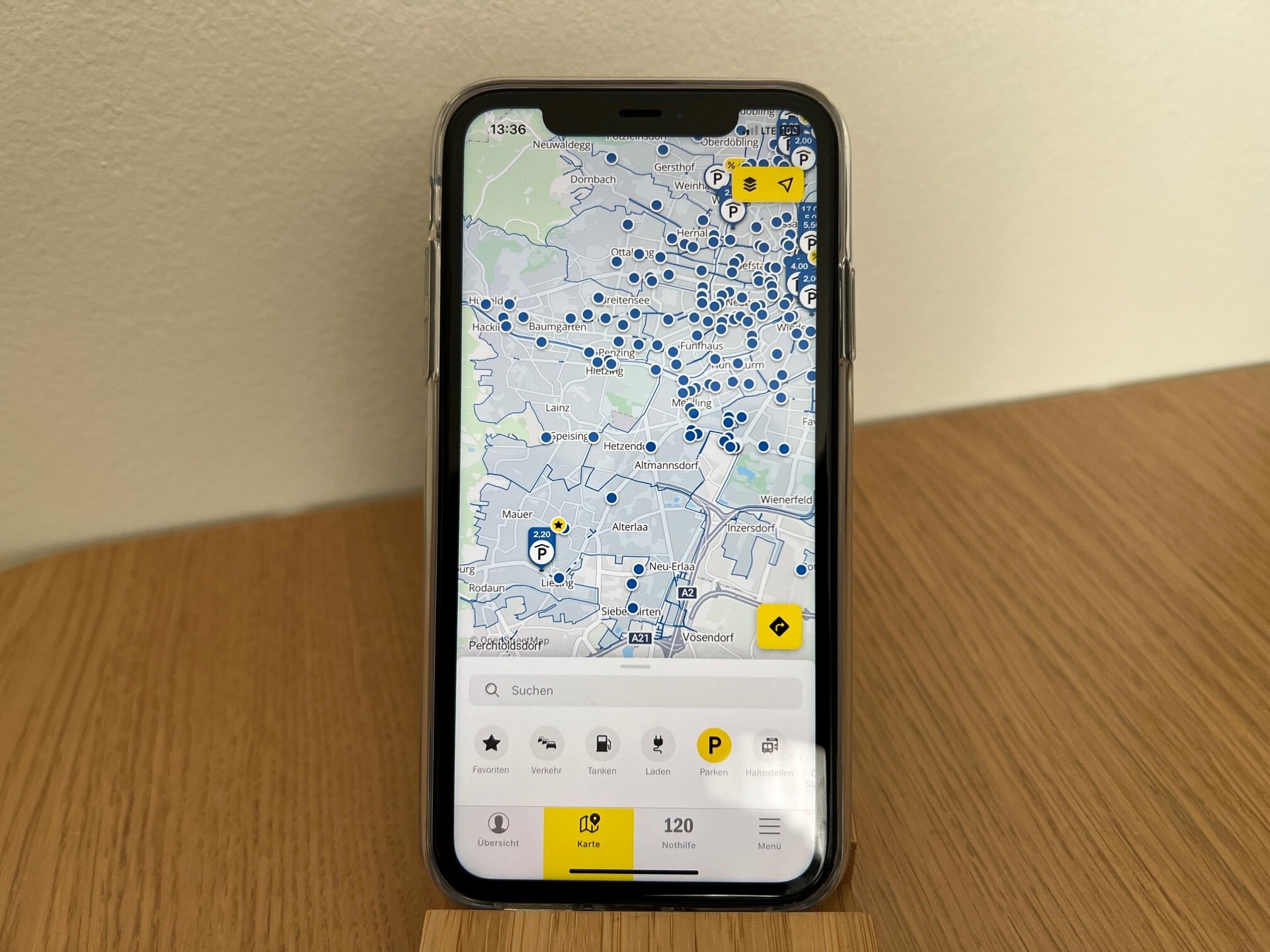 Das Bild zeigt ein Smartphone, das auf einem Holztisch steht. Auf dem Bildschirm ist eine Kartenanwendung der ÖAMTC App geöffnet, die mit blauen Markierungen Parkmöglichkeiten anzeigt.