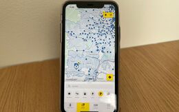 Das Bild zeigt ein Smartphone, das auf einem Holztisch steht. Auf dem Bildschirm ist eine Kartenanwendung der ÖAMTC App geöffnet, die mit blauen Markierungen Parkmöglichkeiten anzeigt.