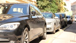 Parkende Autos stehen am Straßenrand einer Kurzparkzone im Sonnenschein. Im Hintergrund sind Mietshäuser, ein großer Baum und ein Kran sichtbar.