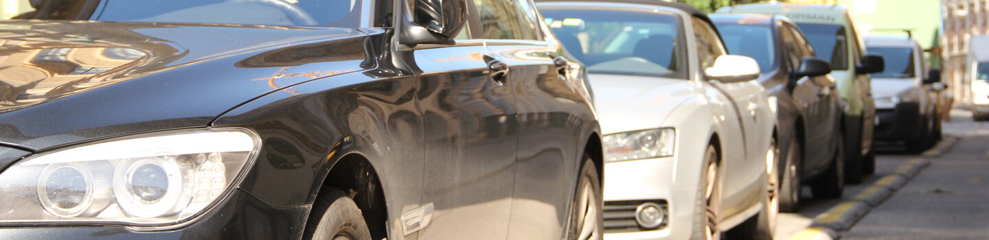Parkende Autos stehen am Straßenrand einer Kurzparkzone im Sonnenschein. Im Hintergrund sind Mietshäuser, ein großer Baum und ein Kran sichtbar.
