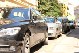Parkende Autos stehen am Straßenrand einer Kurzparkzone im Sonnenschein. Im Hintergrund sind Mietshäuser, ein großer Baum und ein Kran sichtbar.