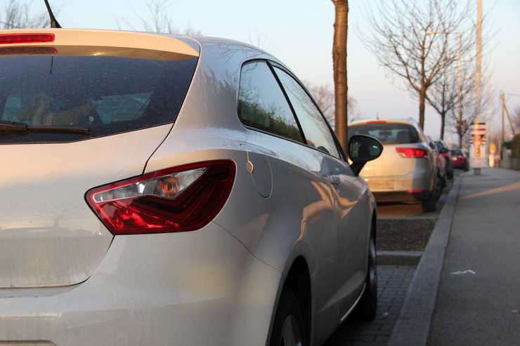 Mehrere Autos parken am Straßenrand, dazwischen sind kleine Grünflächen mit einem Baum.