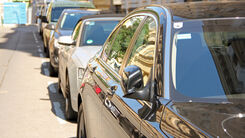 Mehrere Autos parken am Straßenrand im Sonnenschein. Im Hintergrund sind Häuser sichtbar.