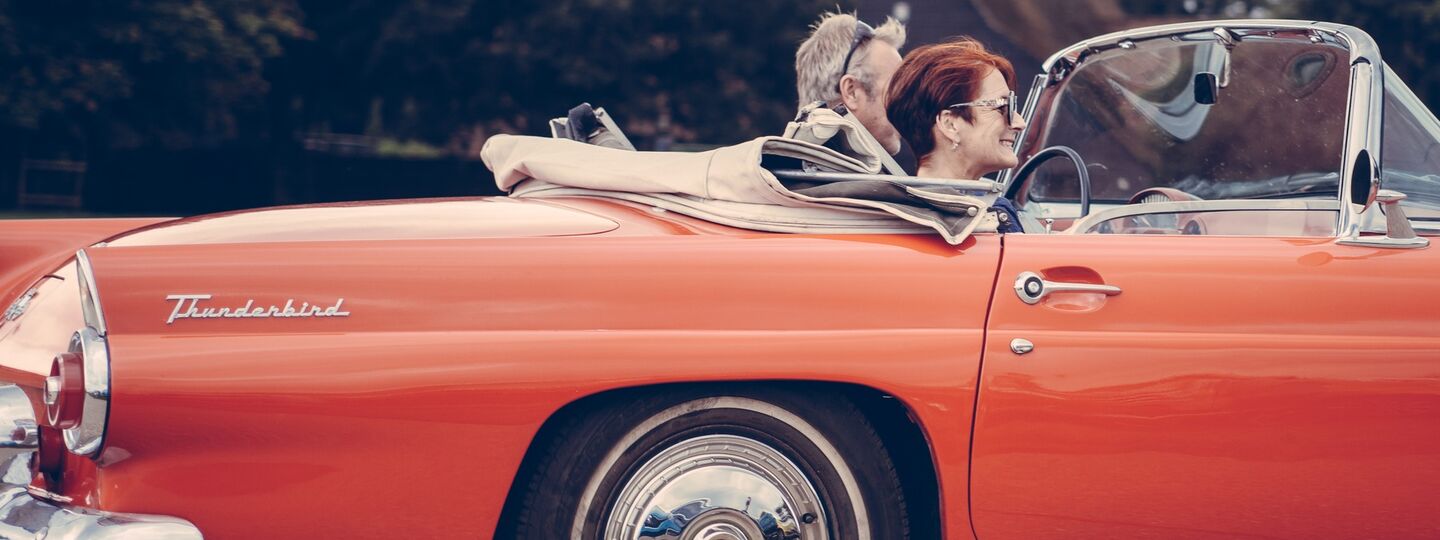 Ein roter Ford Thunderbird Oldtimer, Kabrio mit offenem Verdeck, die Motorhaube ist abgeschnitten. Eine Frau mit kurzen roten Haaren sitzt am Beifahrersitz, ein Mann mit kurzen grauen Haaren fährt das Auto. Im Hintergrund sind Bäume.