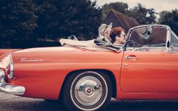 Ein roter Ford Thunderbird Oldtimer, Kabrio mit offenem Verdeck, die Motorhaube ist abgeschnitten. Eine Frau mit kurzen roten Haaren sitzt am Beifahrersitz, ein Mann mit kurzen grauen Haaren fährt das Auto. Im Hintergrund sind Bäume.