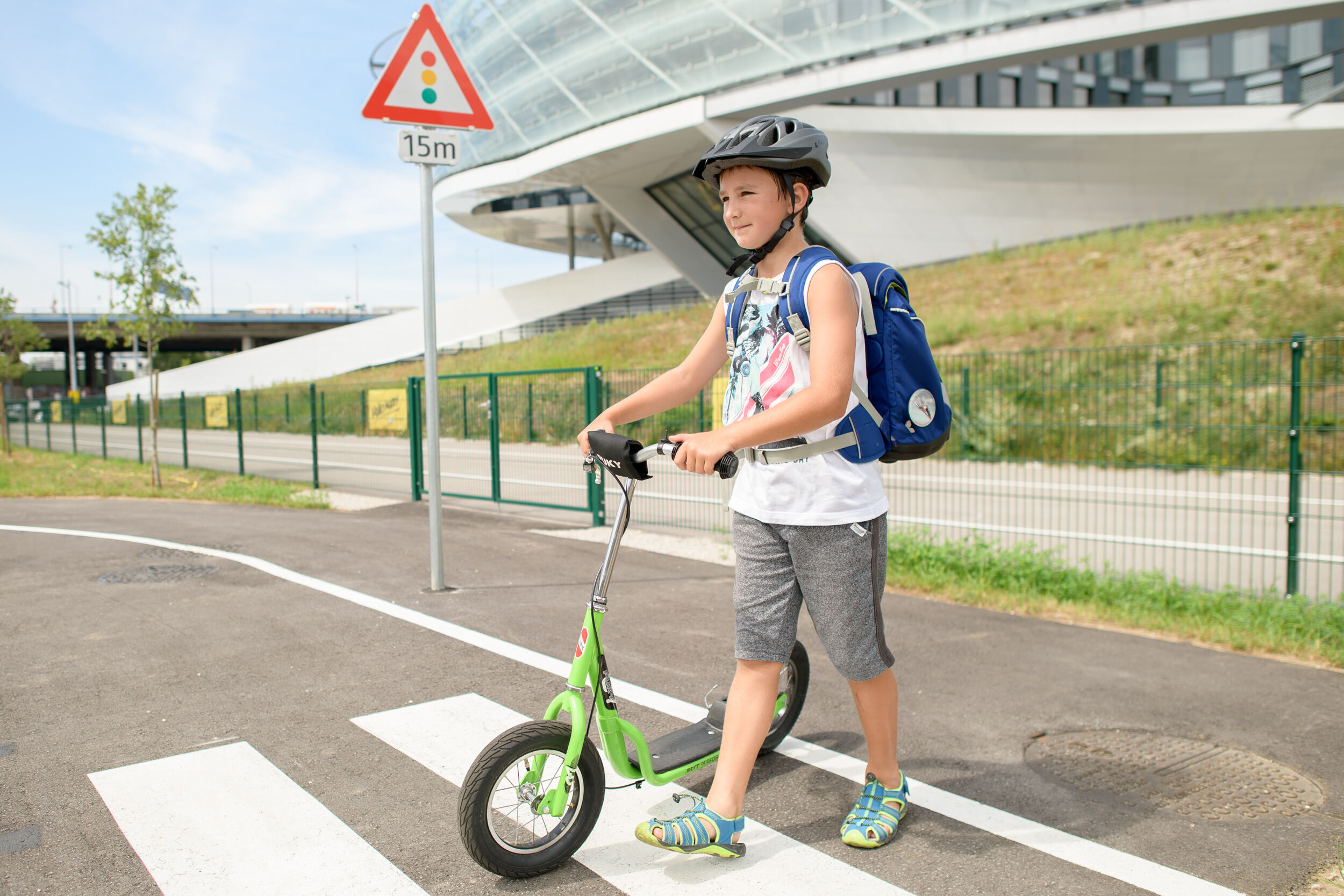 Ein Schulkind mit Schultasche am Rücken und Fahrradhelm am Kopf schiebt einen grünen Scooter über einen Zebrastreifen. Im Hintergrund sind ein asphaltierter Weg und ein großes Bürogebäude.