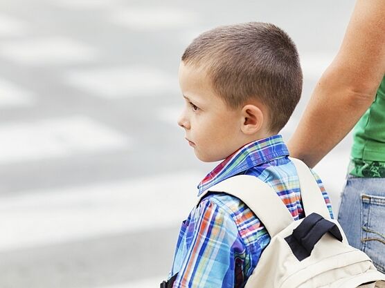Ein Schulkind mit Rucksack steht mit einer Begleitperson an einem Zebrastreifen und blickt auf die Straße.