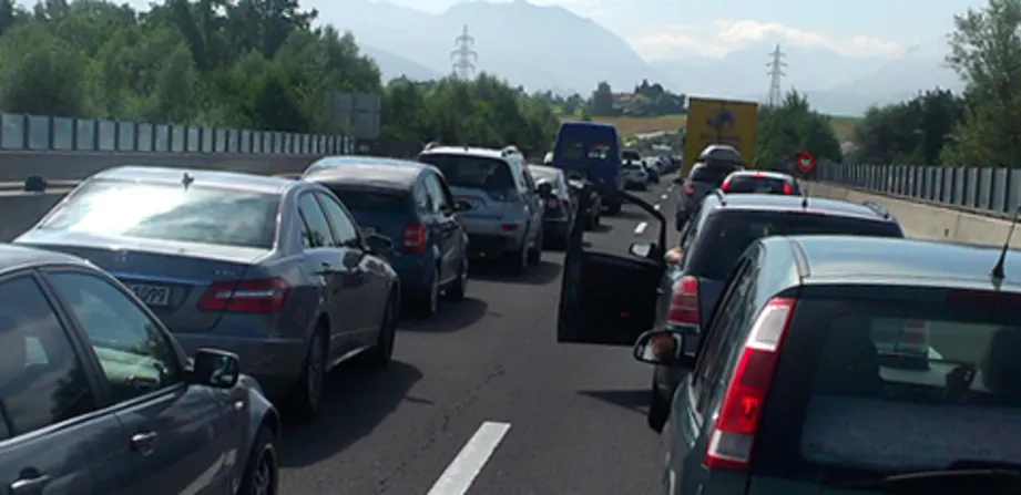 Stau auf einer Autobahn, viele Autos stehen auf beiden Fahrspuren in der Sonne. Im Hintergrund Landschaft mit Bäumen.
