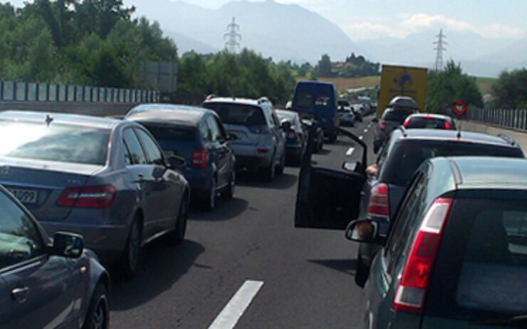 Stau auf einer Autobahn, viele Autos stehen auf beiden Fahrspuren in der Sonne. Im Hintergrund Landschaft mit Bäumen.