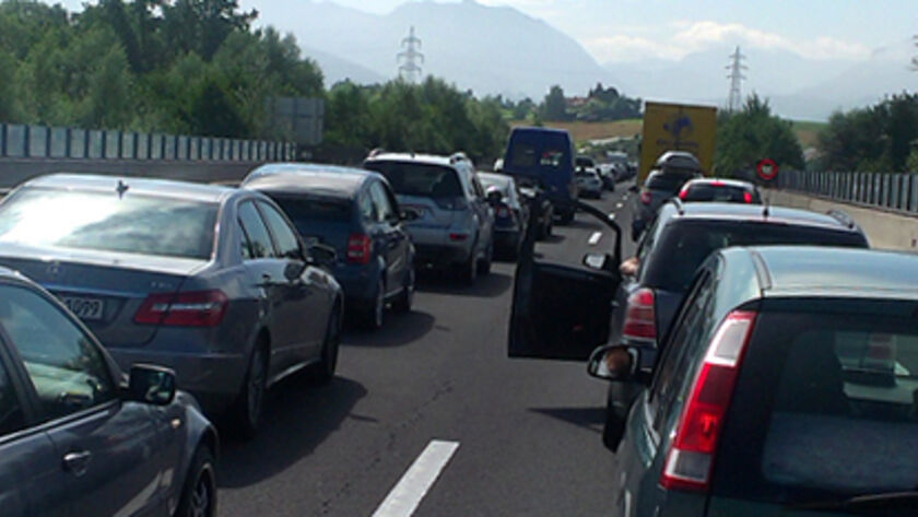 Stau auf einer Autobahn, viele Autos stehen auf beiden Fahrspuren in der Sonne. Im Hintergrund Landschaft mit Bäumen.