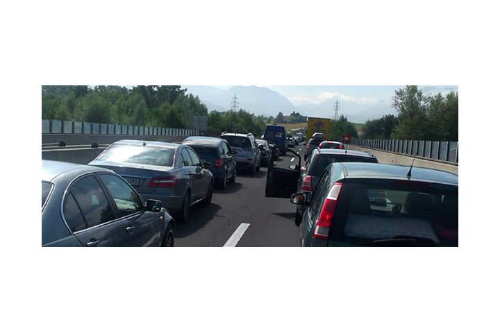 Stau auf einer Autobahn, viele Autos stehen auf beiden Fahrspuren in der Sonne. Im Hintergrund Landschaft mit Bäumen.
