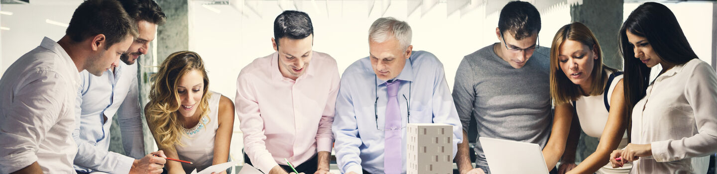 In einem Besprechungsraum stehen an einem Tisch fünf Männer und drei Frauen. Am Tisch liegen große Papierpläne und ein Architekturmodell von einem Gebäude. Die Gruppe bespricht die Pläne.