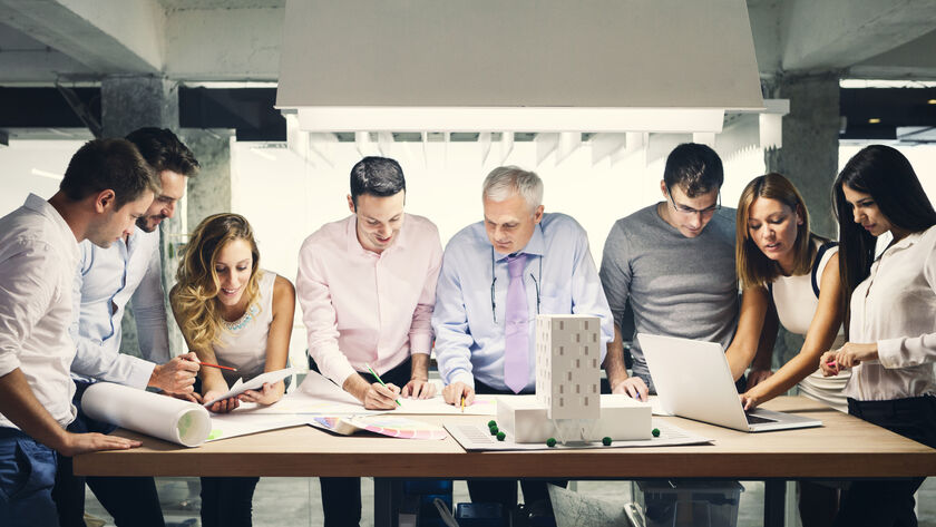 In einem Besprechungsraum stehen an einem Tisch fünf Männer und drei Frauen. Am Tisch liegen große Papierpläne und ein Architekturmodell von einem Gebäude. Die Gruppe bespricht die Pläne.
