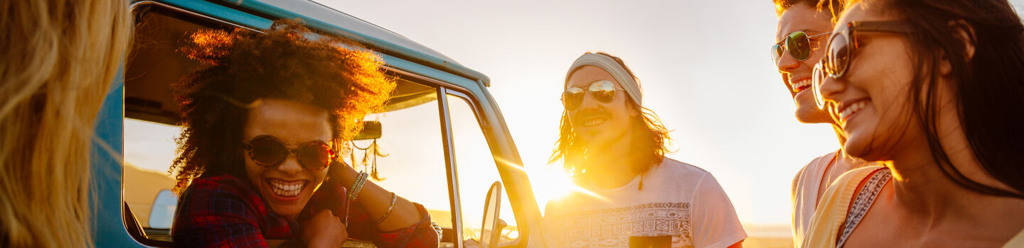 Im Sonnenuntergang steht auf einer Wiese ein alter, türkiser Bus, aus dem Fenster lehnt sich eine Frau mit Sonnenbrille und lacht. Neben dem Bus stehen zwei Männer und zwei Frauen, alle lachen, es ist eine entspannte Stimmung.