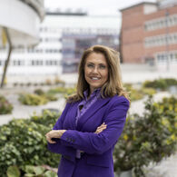 Seidenberger Marion Auf dem Bild sieht man eine Dame mit verschränkten Armen. Sie trägt einen violetten Blazer und eine schwarze Hose. Sie schaut in die Kamera und lächelt. Im Hintegrund sieht man grüne Pflanzen.