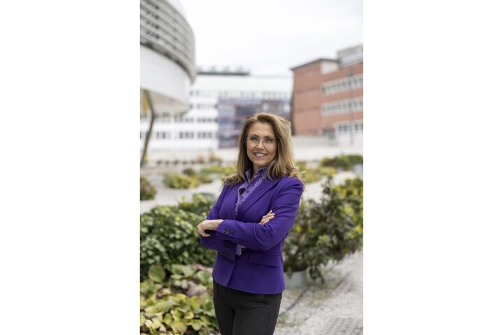 Auf dem Bild sieht man eine Dame mit verschränkten Armen. Sie trägt einen violetten Blazer und eine schwarze Hose. Sie schaut in die Kamera und lächelt. Im Hintegrund sieht man grüne Pflanzen.